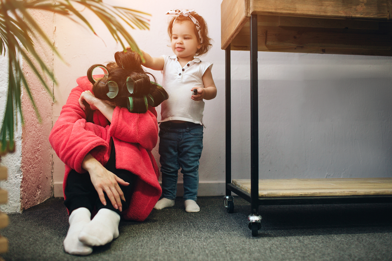 New mother in robe and rollers sitting on the floor with head in arms while young daughter tries to comfort her. Postpartum help is available in Tigard, OR 97223