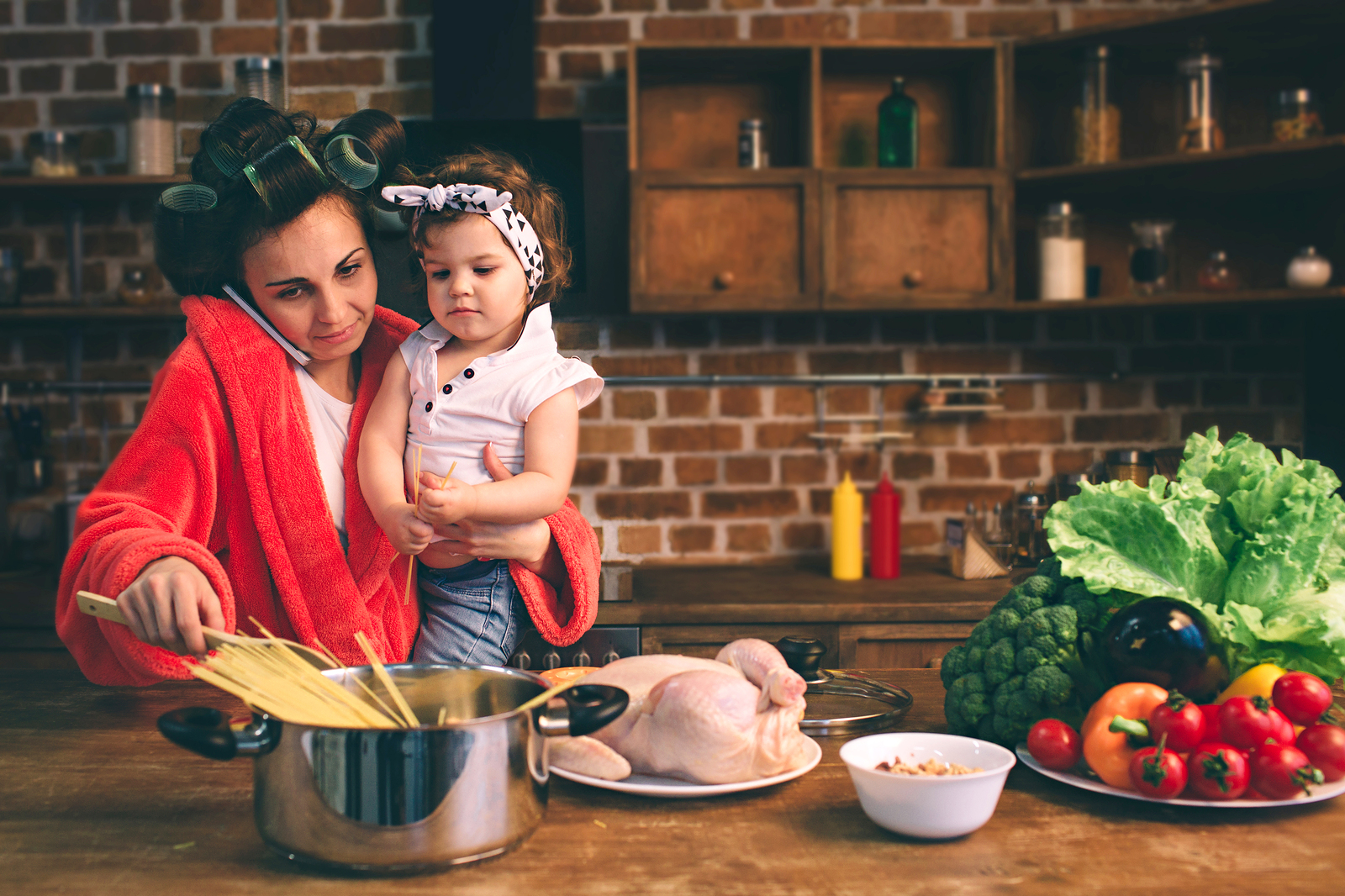 Mom cooking dinner with robe on, curlers in hair, talking on phone while holding a baby in her kitchen. Postpartum parents need counseling support in Tigard Oregon