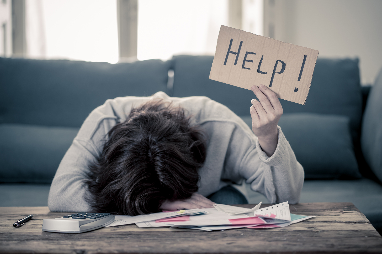 Person with head down on a desk covered in papers, looking distraught. Holding a paper, handmade sign that says "Help!". This person could definitely benefit from Portland-area counseling for life transitions. 